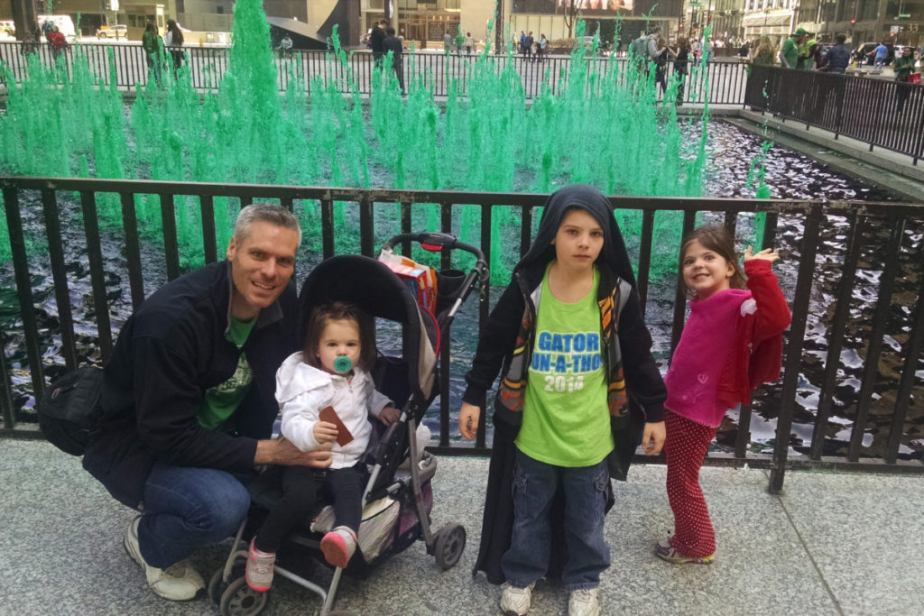 A man and 3 little children stand in front of a fountain dyed green for St. Patrick's Day