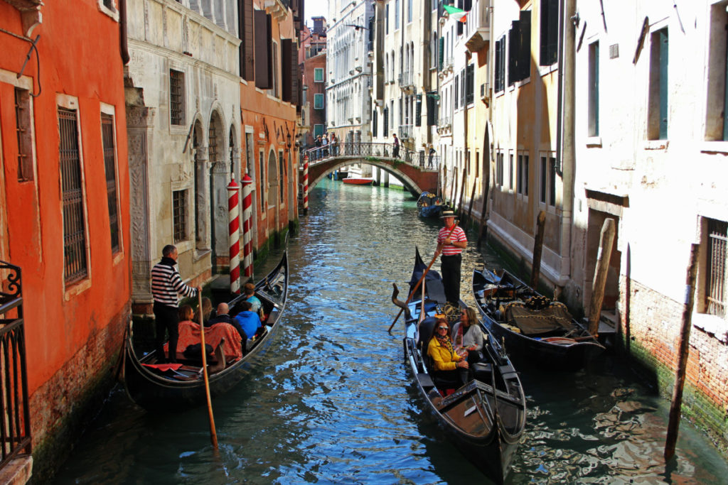 a man guides a gondola in Venice, Italy with a bridge and bright buildings in the background