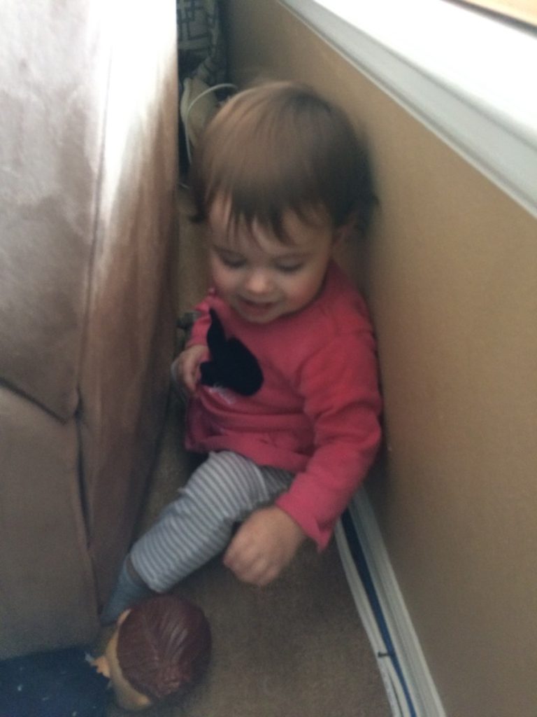 A toddler hides behind the couch from mom
