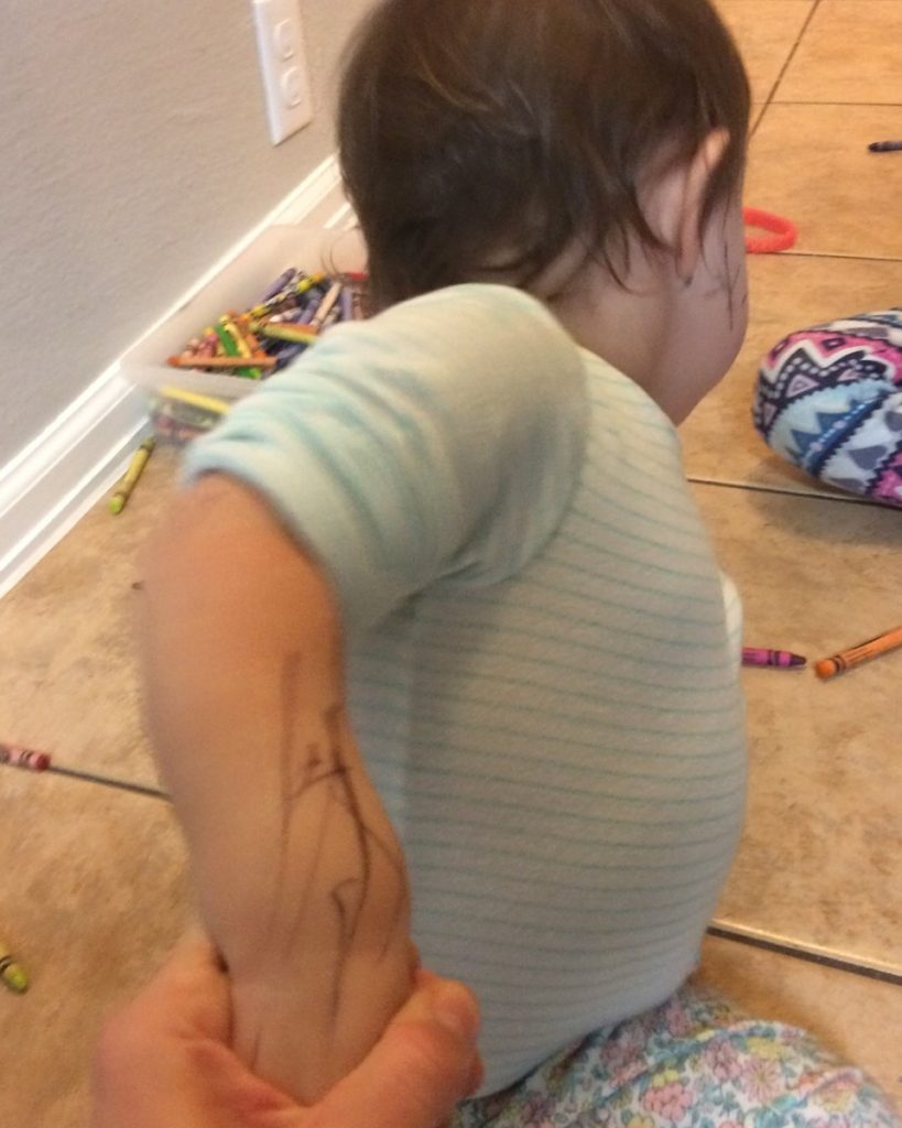 A toddler draws on her arm with a pen