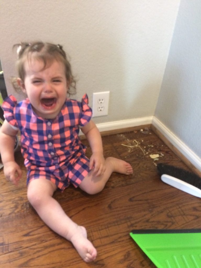 A toddler cries next to a pile of dirt and a broom and dust pan