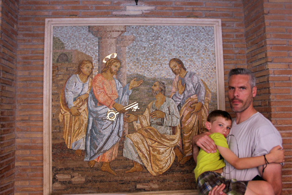 Father holds a boy outside St. Peter's basilica with a mosaic of Jesus giving keys to His apostles. 