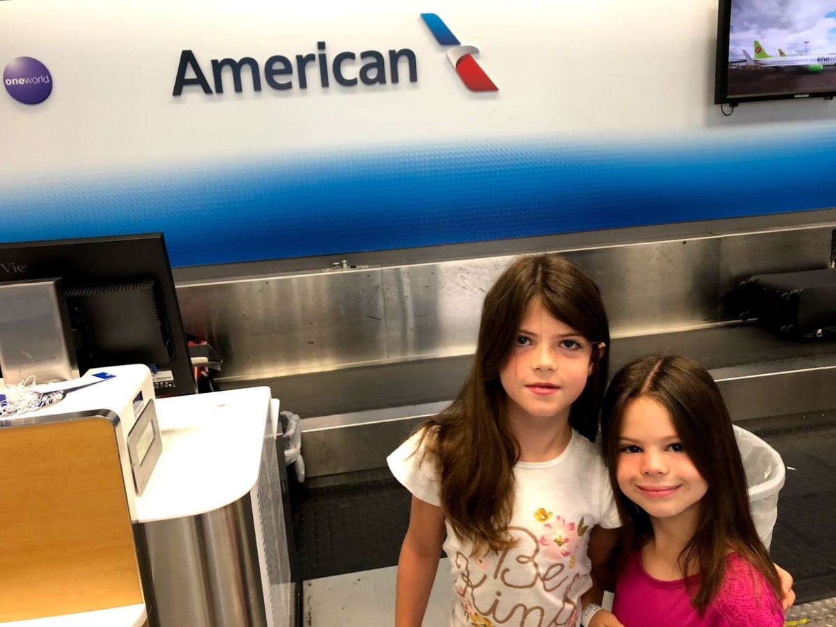 2 girls smile in from of baggage carosel at American Airlines counter