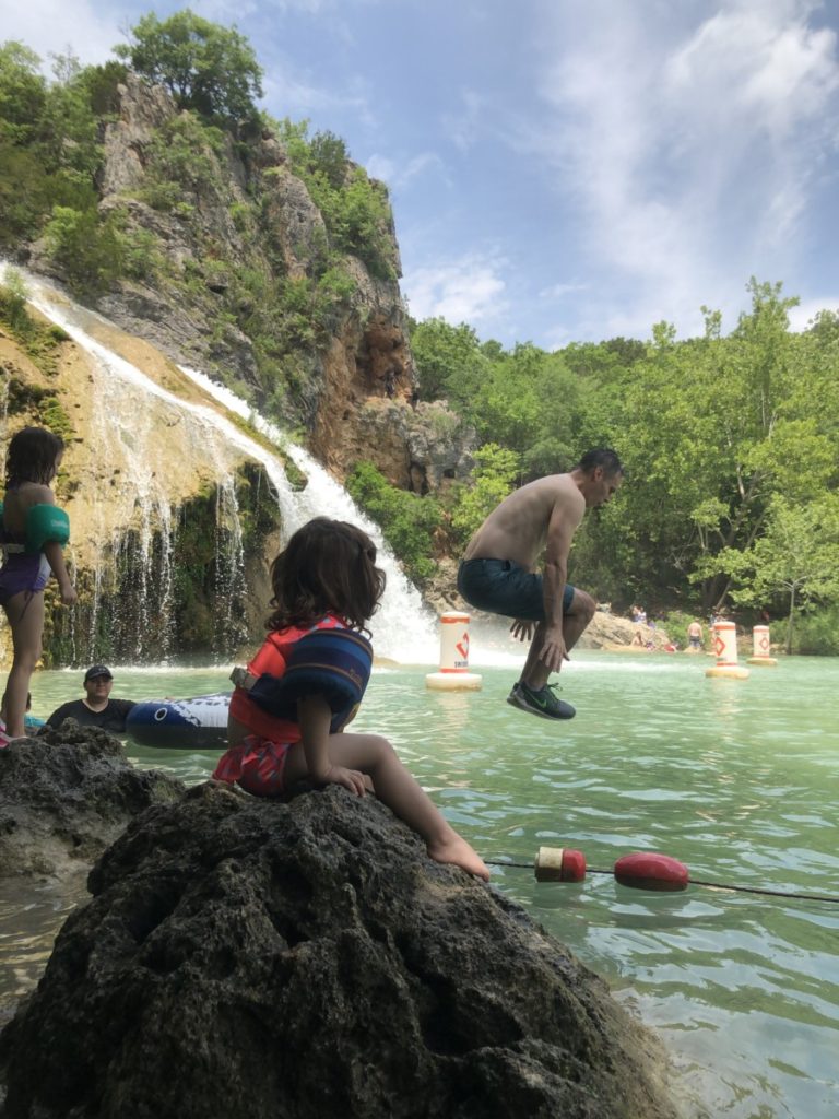 A toddler watches from a rock as her dad jumps into the water below Turner Falls