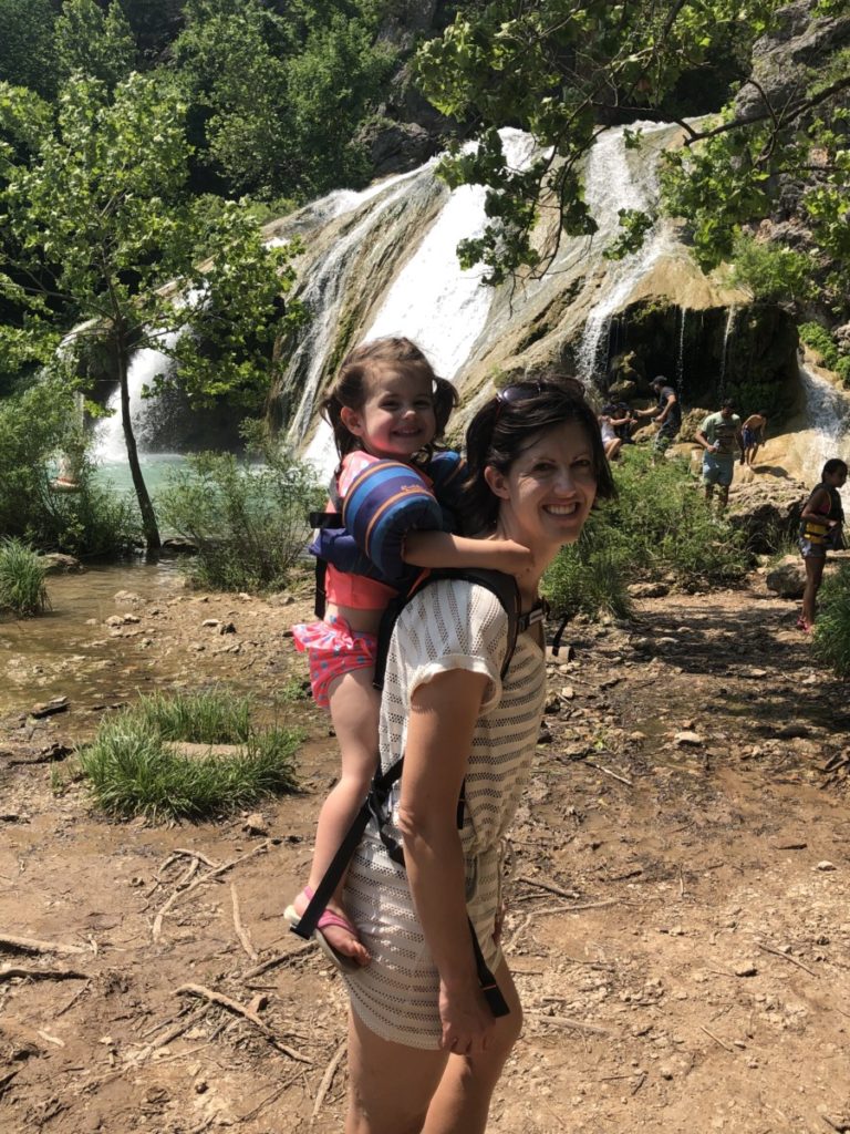 A toddler stands on the Piggyback Rider with Turner Falls in the Background
