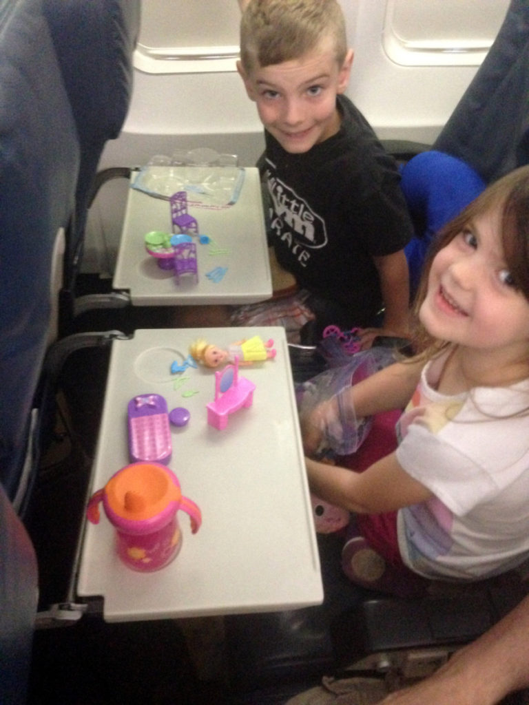 A brother and sister play with doll furniture on the tray tables of an airplane