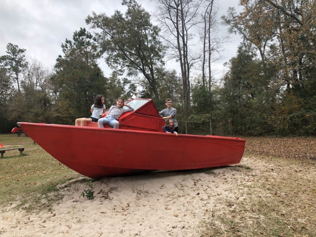 3 kids sit on a red boat that is beached at 7 acre Wood in Conroe Texas