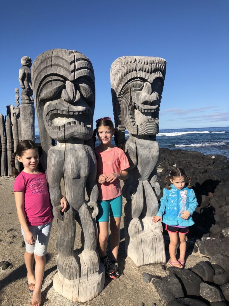 3 girls stand with 2 hawaiian statues