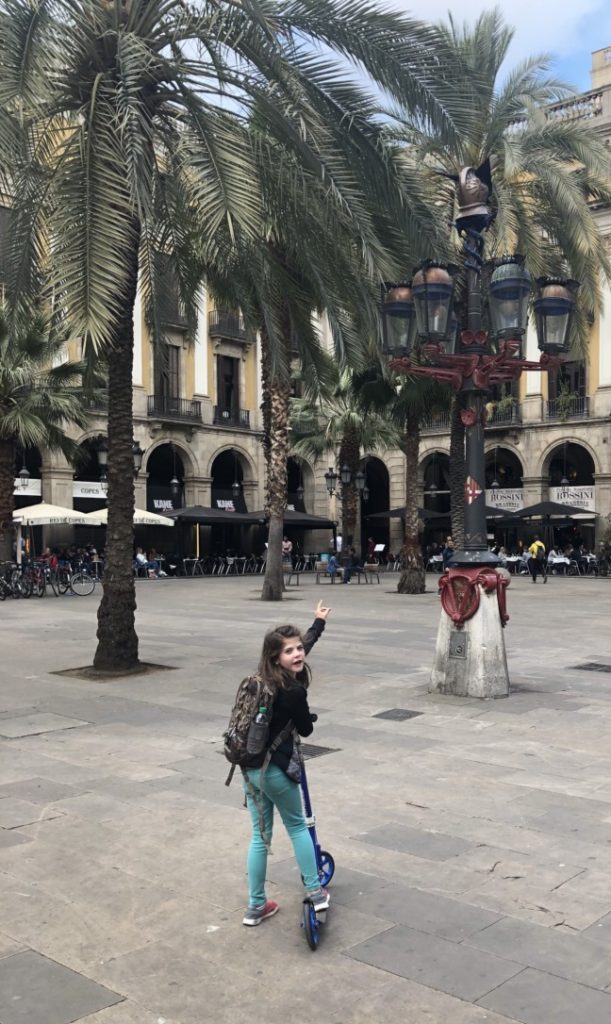 A girl pointing to a lamp pole while riding a scooter through Plaza Reial in Barcelona