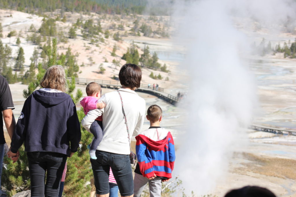 Grandparents and mom with 2 kids walking through Yellowstone National Park Norris Geyer Basin with steam in the background
