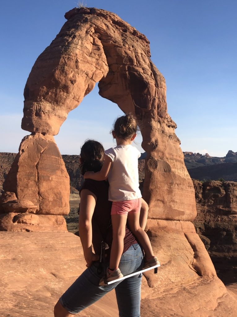 Mom holds daughter on her back in the Piggy back riders at delicate arch in Arches National Park