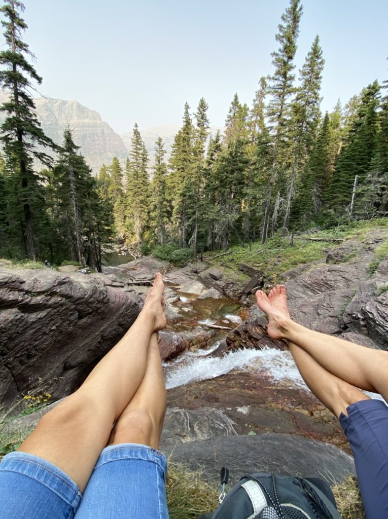 Foot selfie with the trees and peaks and waterfalls of Glacier National Park in the Distance