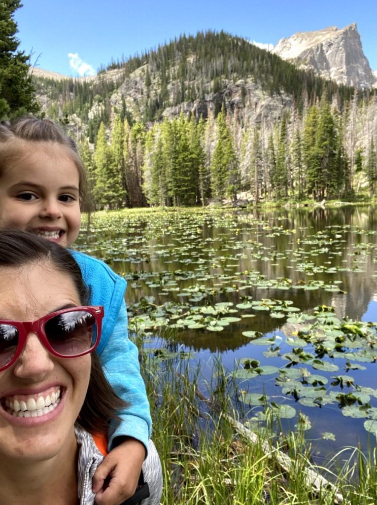 mom hiking with kids.  Toddler on the piggyback rider on her back. Nymph Lake with lily pads in Rocky Mountain National Pakr