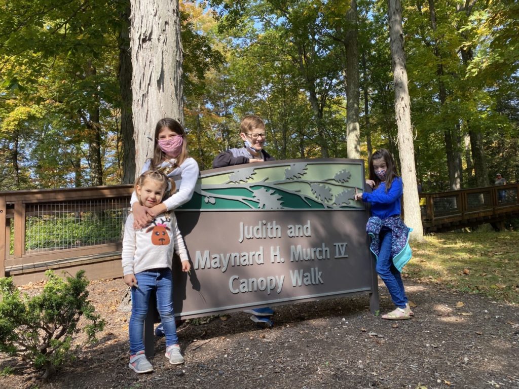 4 kids stand in masks near the sign of the Judith and Maynard Murch Canopy Walk in the Holden Arboretum