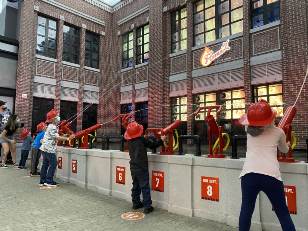 6 kids in firefighter hats shoot water at a burning building in Kidzania in Frisco, Texas
