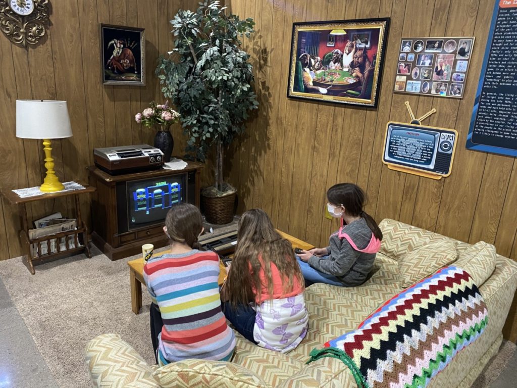 3 kids sit on a retro couch and room and play old video games like frogger at the National Video Game Museum in Frisco